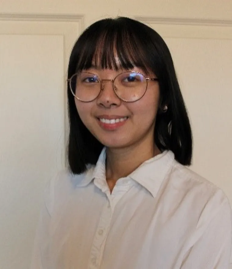 Nadine, an Asian person with shoulder-length black hair and bangs, is smiling in front of a white background. They are wearing gold-rimmed glasses and a white button-down shirt.