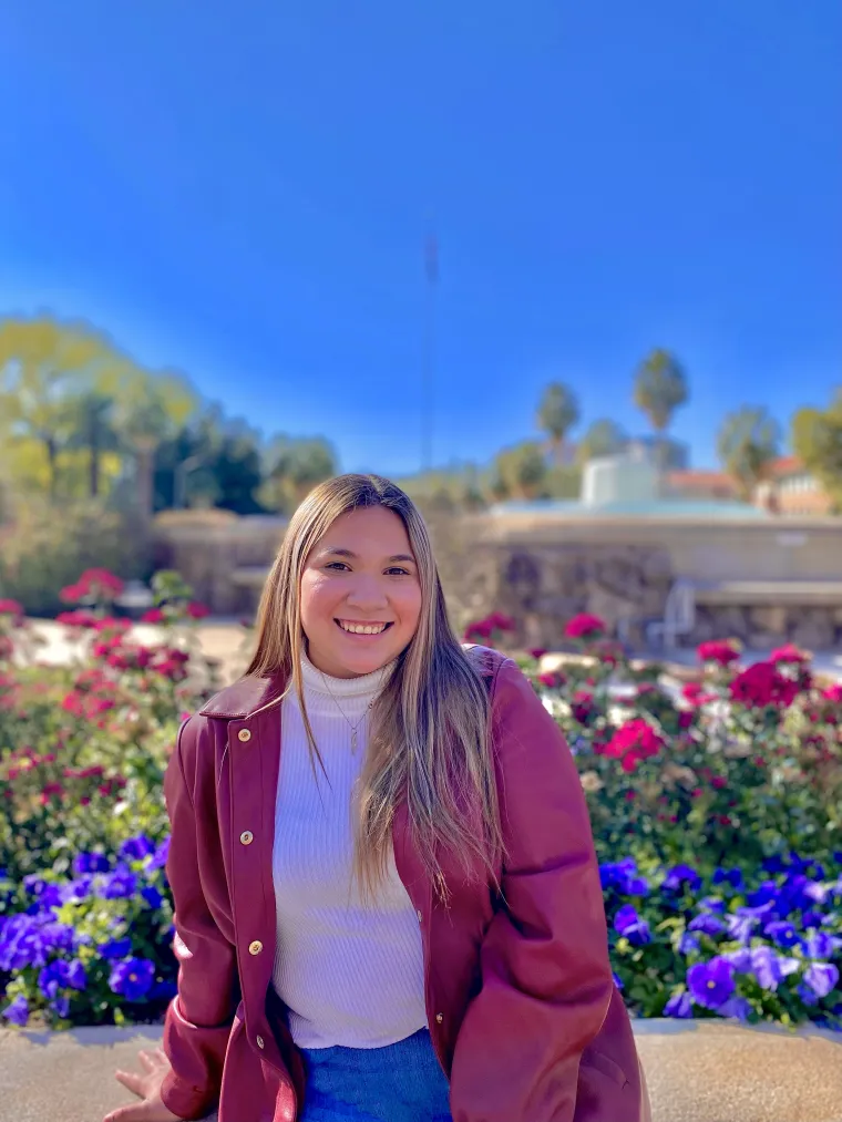 Paulette is wearing a white turtle neck shirt with a red jacket and jeans. Paulette's hair is blonde with brown roots, she is light-skinned and is smiling. Paulette is sitting down in front of a water fountain with flowers in the background during a sunny day.