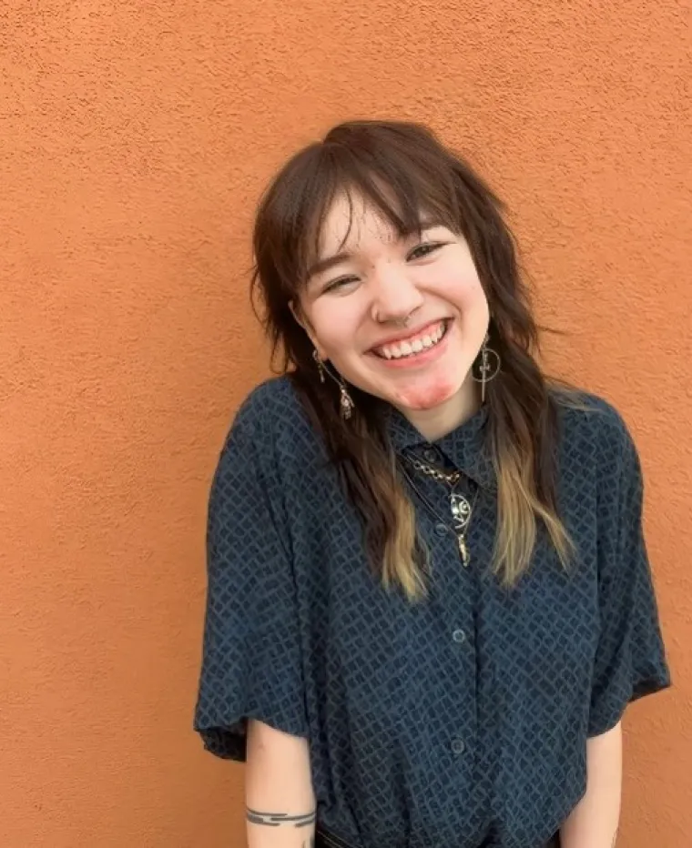 Ree, a Chinese American individual with blue and black checkered button up shirt. They are smiling in front of an orange wall outside on a bright day, wearing long alternative earrings, 3 necklaces, and their hands in their pockets.