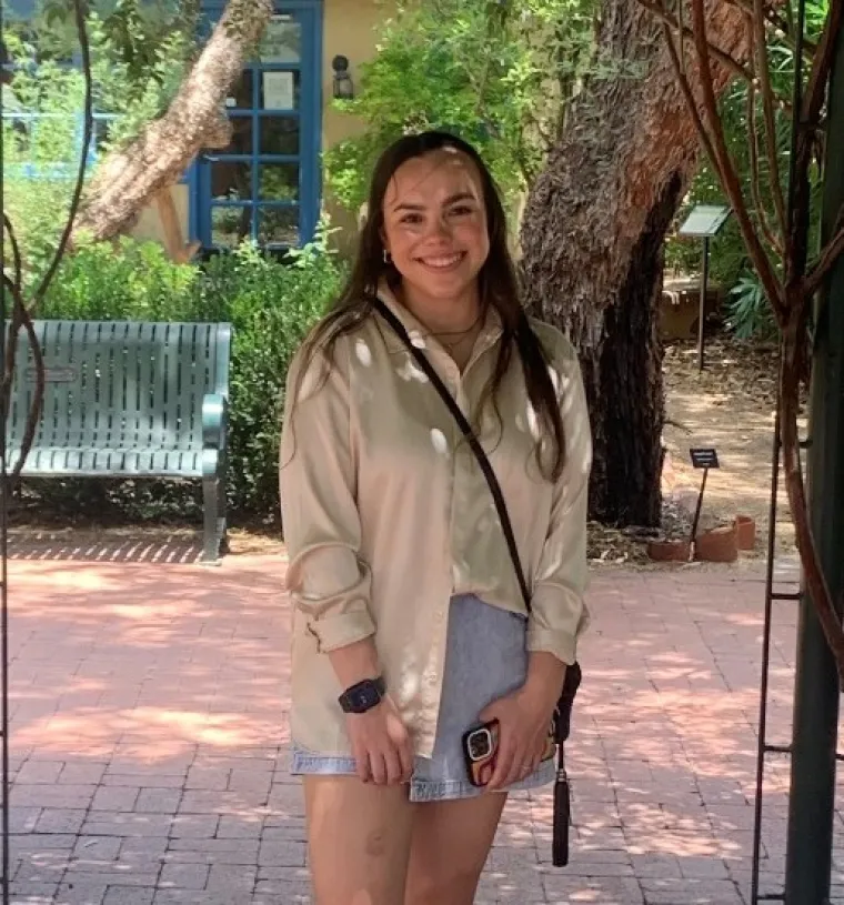 In this image, I am wearing a gold satin blouse with a denim skirt. My hair is down and I am also wearing a black small purse. The background of the image shows trees and plants from the Tucson Botanical Garden.