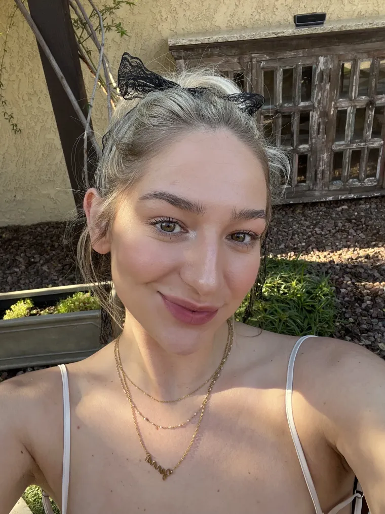 I am a white female, with blonde hair and a black bow in my hair. I am wearing a gold necklace with my name on it and a cream colored dress. Smiling at the camera