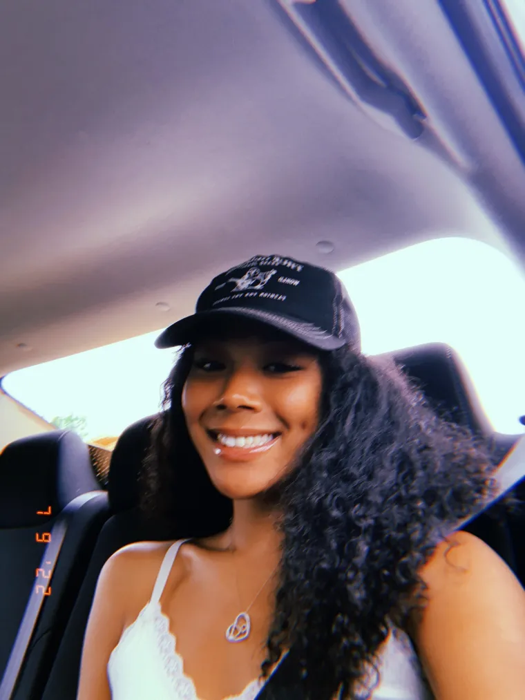 Brown skinned female with long curly hair selfie taken in the back of a car, wearing a black/white hat, white tank top, with a silver/gold necklace, smiling at the camera