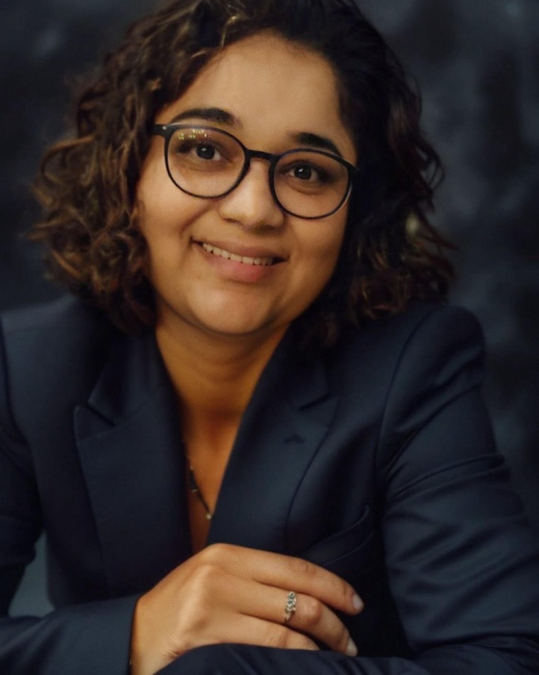 Woman with short, curly dark hair wearing glasses and a dark grey blazer.