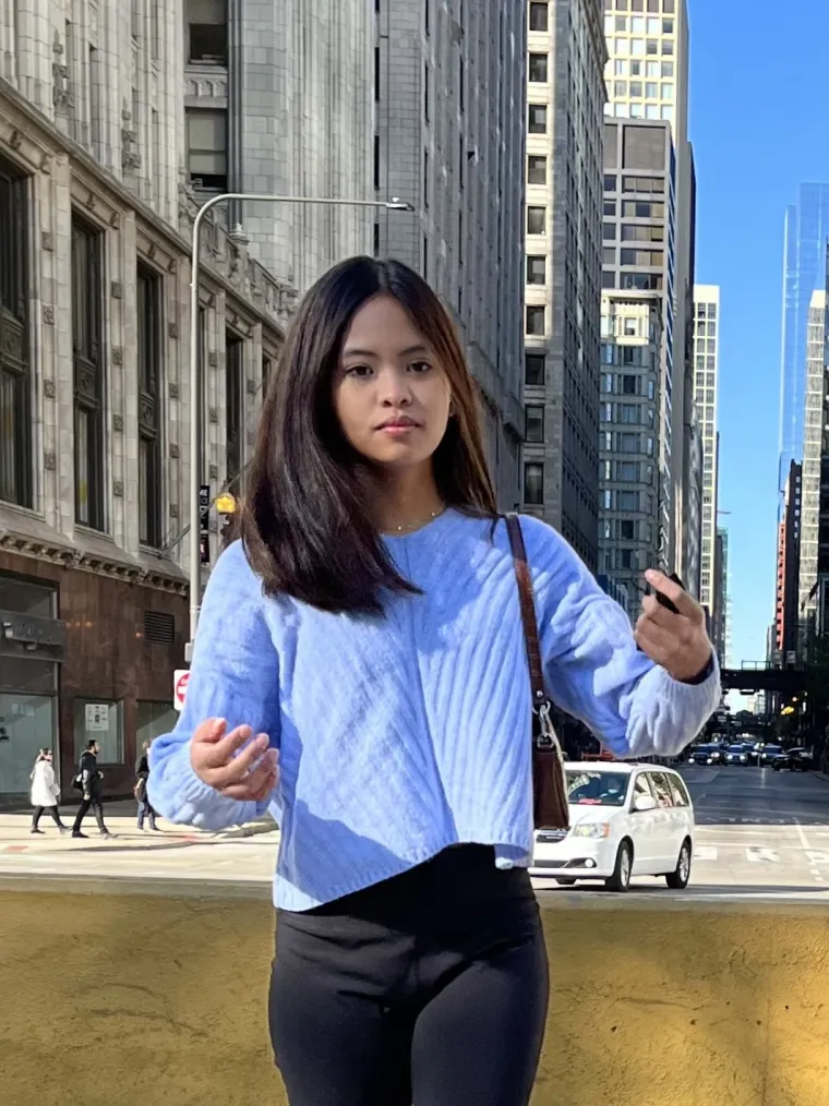 Dana has shoulder length brunette hair, standing in front of buildings wearing blue sweater look at the camera. 