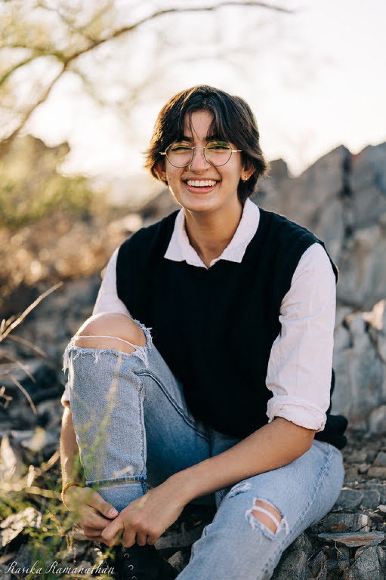 student sitting on rocks in front of desert background, wearing a white collared shirt with lack vest and jeans, swearing glasses with short brunette hair smiling at the camera