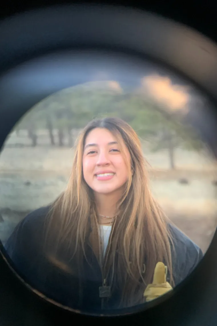 Anahy, a hispanic woman with long brown hair, is outside. She wears a black jacket and smiles at the camera.