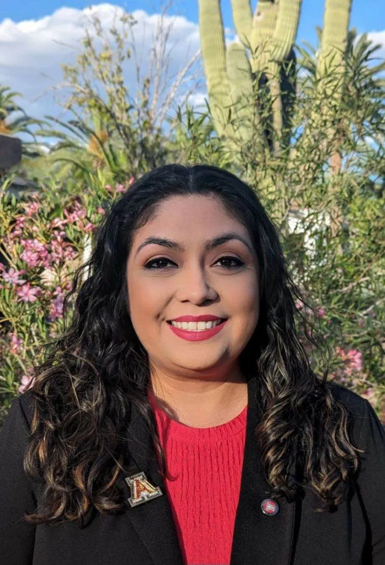 young woman smiling at camera, there are cacti and flowers in the distance. She is wearing a red shirt with a black blazer and a University of Arizona logo pinned on top. 