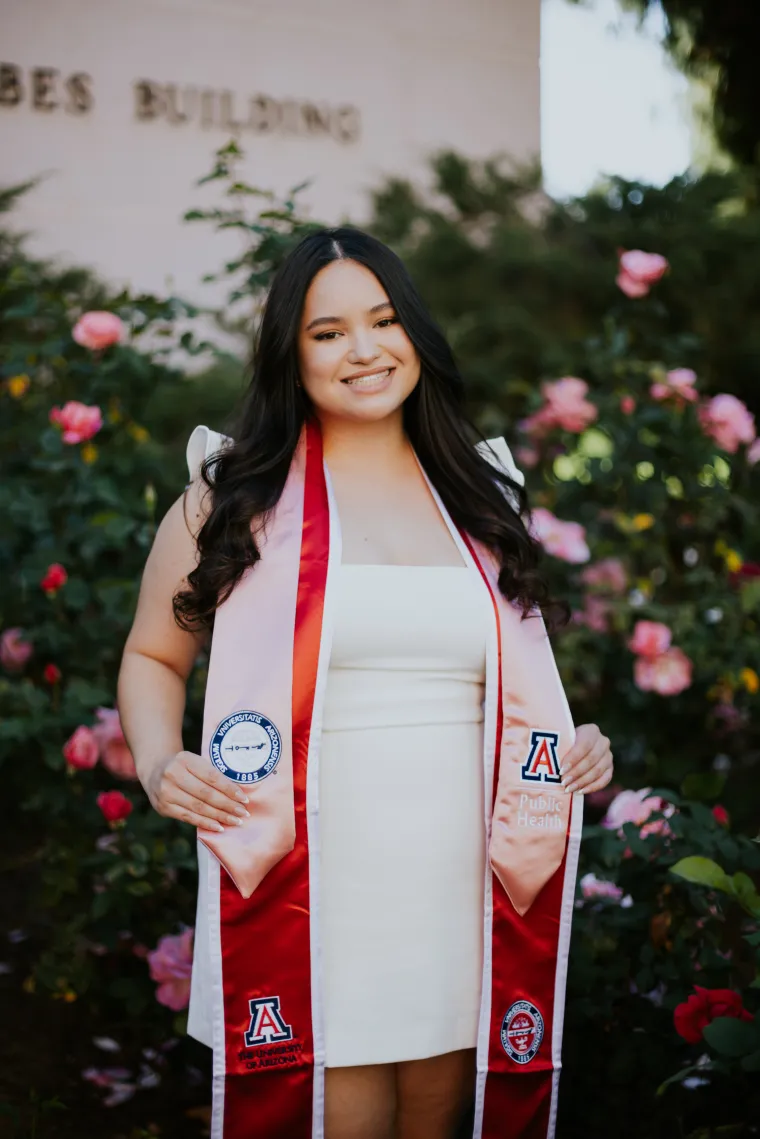 Photo of young woman in her University of Arizona cap and gown, long black hair smiling at camera. Standing in front of rose bushes