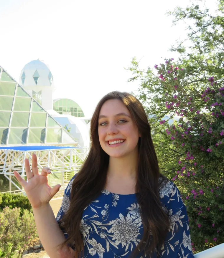 Nicole, a white woman with long brown hair, is outside on a bright day. The background includes a green bush with small purple flowers, and mountains in the background. She leans her side against a railing and smiles at the camera. She wears a royal blue blouse with white flowers, and black dress pants.