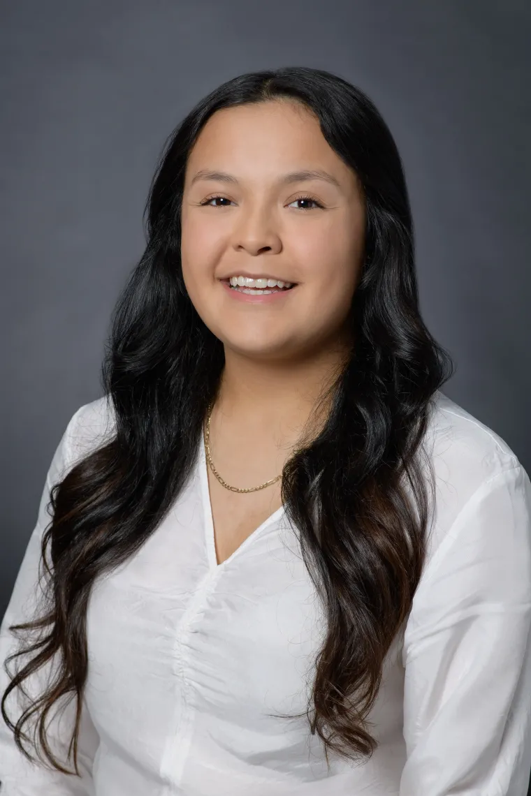 A young woman in front of a grey backdrop, with long brunette hair and wearing a white button down shirt smiling at the camera