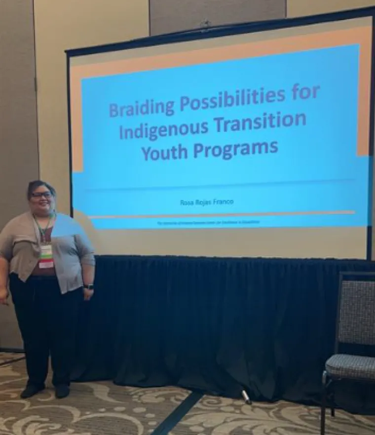 Rosa Rojas-Franco presents on Indigenous Youth Transition Programs at the APSE conference. She is standing in front of a powerpoint slide reading "Braiding Possibilities for Indigenous Transition Youth Programs", and smiling.