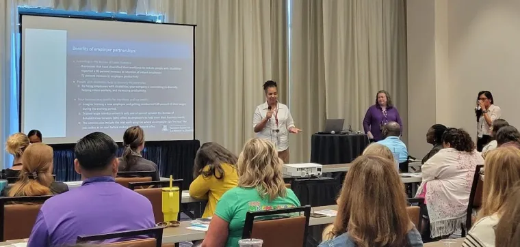 Lisa Ayo and Kelsey Montaño presents with Traci Przecioski from AZ Vocational Rehabilitation on Pre-Employment Transitions Services and the benefits of employer partnerships. Lisa is standing, speaking into a microphone in front of a crowd of attendees. Behind her is a powerpoint slide that reads "benefits of employer partnerships"