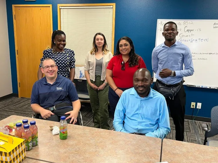 Denis Ouma (2nd from right) and support staff Stella (right) and Allan (2nd from left) visit Jason Snead (left) at the Arizona Developmental Disabilities Planning Council, accompanied by the Sonoran Center’s Ava Gutierrez and Celina Urquidez (center left, center right). All are gathered indoors behind a table with some refreshments on it.