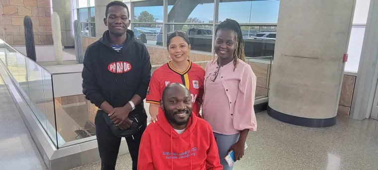 Denis Ouma seated in his wheelchair at Tucson International Airport. Sonoran Center director Jacy Farkas and support staff Allan and Stella stand behind him. All are smiling.