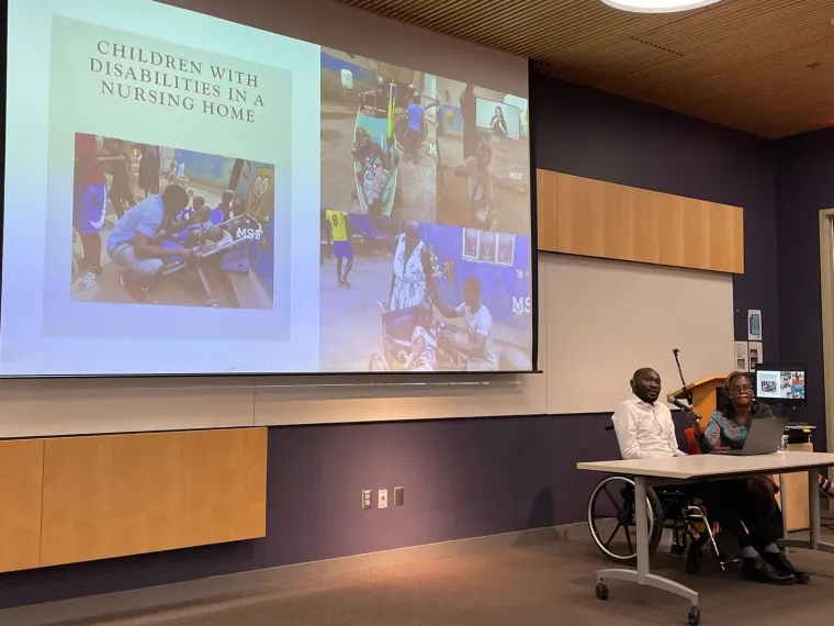 Denis is seated next to a projector displaying the first slide of his presentation on the state of disability supports in Uganda. The slide reads "Children with disabilities in a nursing home" and includes pictures of children doing various activities. A support person is seated next to Denis.