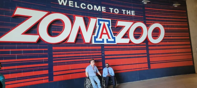 UArizona adaptive athletics director Pete Hughes (left) and Denis (right) at ZonaZoo, the UArizona official student section and ticketing program for UA Athletics. Pete and Denis are posing for the camera beneath a large sign with the words "welcome to the Zona Zoo".