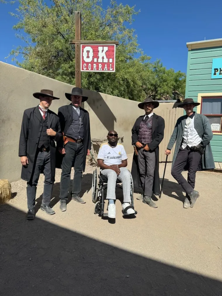 Denis (center) and actors at the OK Corral in Tombstone, AZ. The four actors are wearing Wild West cowboy attire. Denis is on his wheelchair between them, with two actors on his left and two on his right. All are standing outside on sand below a sign which reads "O.k. Corrall". 