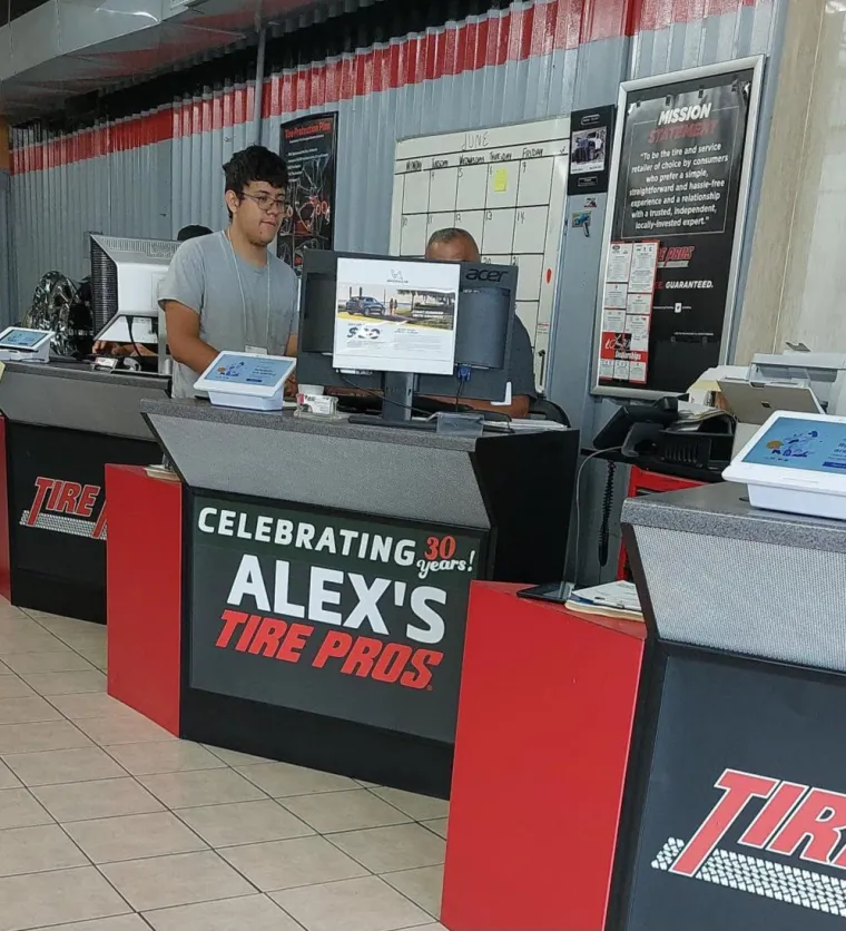 Student worker Nathaniel stands behind the front desk at Alex's Tire Pros, looking at a computer. He is looking at a computer monitor while another employee (face obscured by the monitor) demonstrates something. 