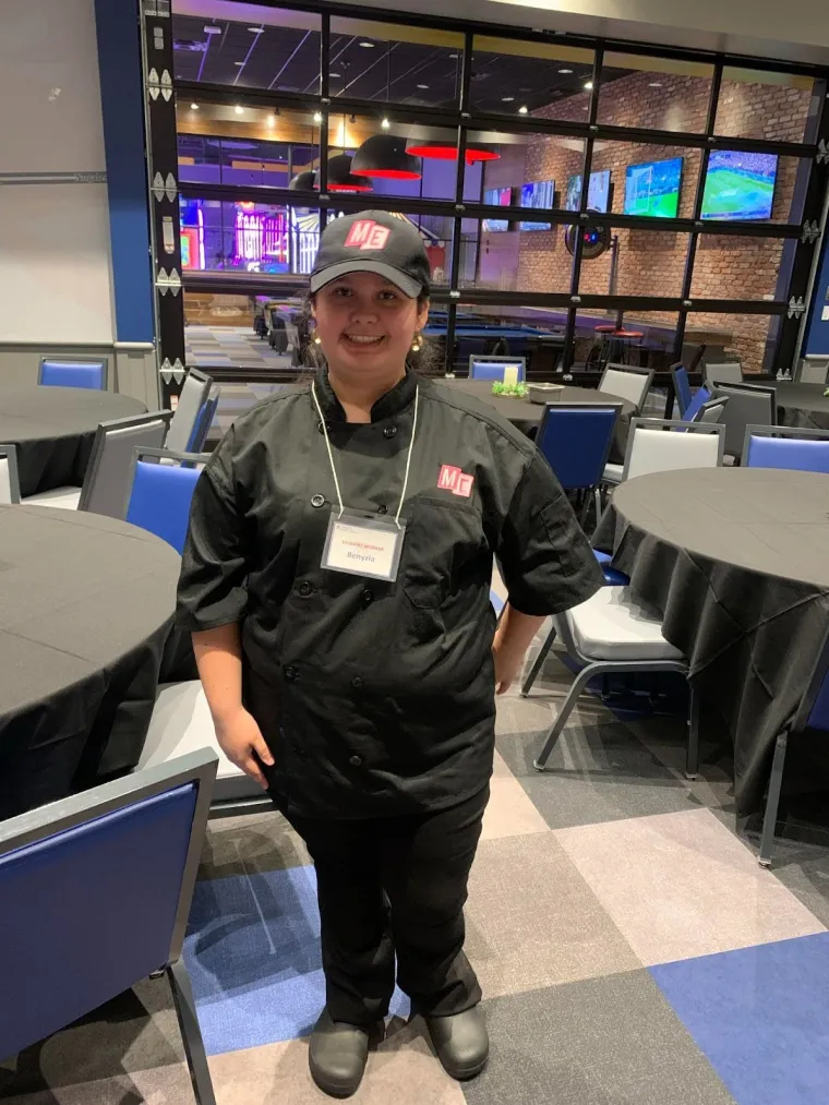 Student worker Benyzia smiles in her uniform at Main Event Tucson. She is wearing a black button up shirt with black pants and shoes, and a black cap with the Main Event logo. Dining tables and pool tables can be seen behind her.