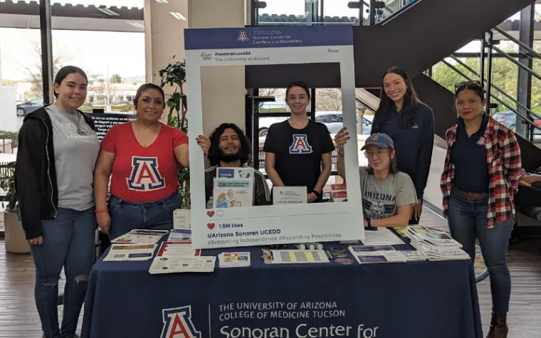 The author (third from right) sits with Sonoran Center staff at a conference table. They are holding a large poster board with a square cut in the middle, and decorated to resemble a social media post. The table has numerous Sonoran Center fliers and brochures on it. 