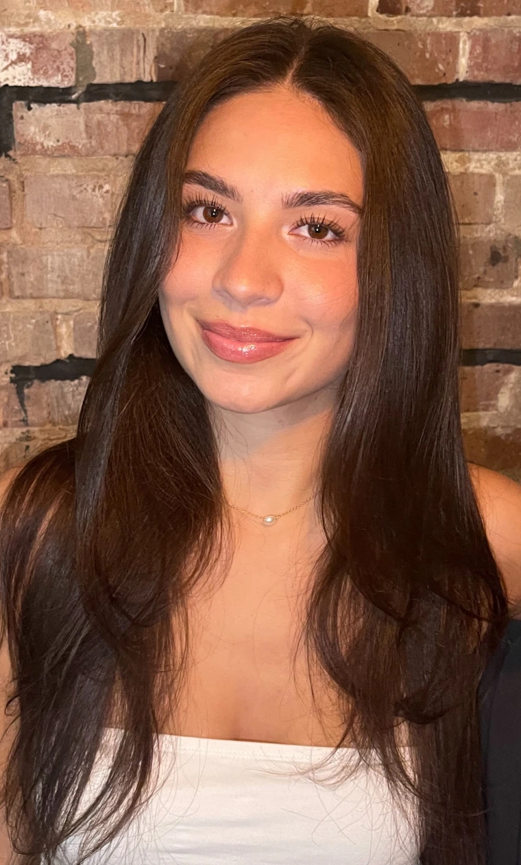 Woman with long brunette hair in front of brick background, smiling at the camera, wearing a white shirt