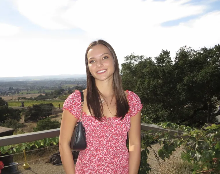 Phot of woman standing in front of green landscape, wearing a pink floral dress with brunette hair is smiling at the camera