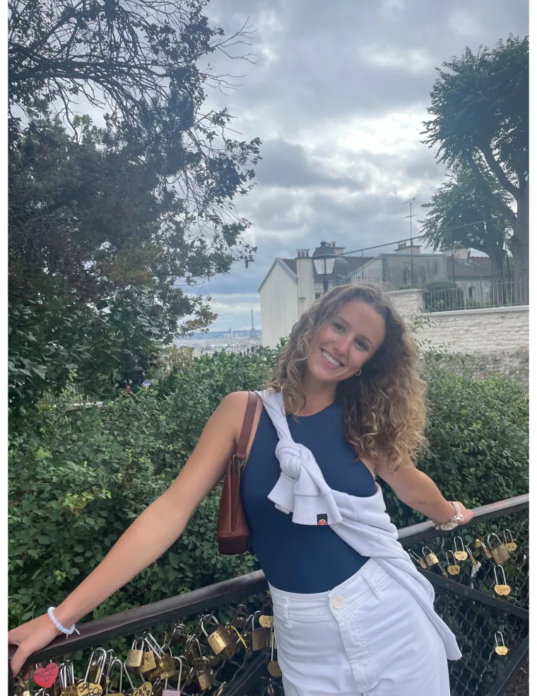 Photo of woman in front of a grassy landscape, wearing a navy blue top and white skirt, with blonde curly hair, smiling at the camera