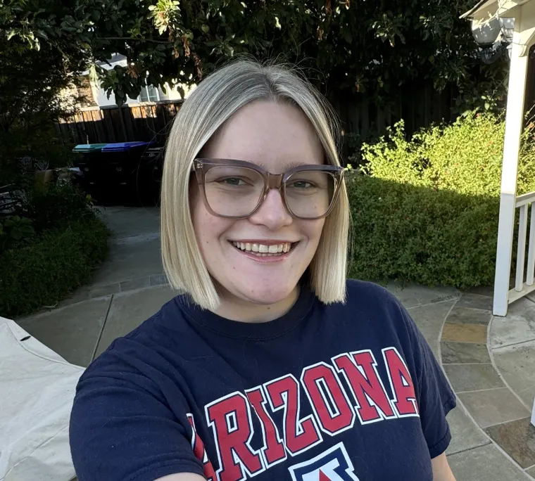 Photo of woman in front of greenery, wearing a University of Arizona shirt. She has a blonde bob and is wearing glasses