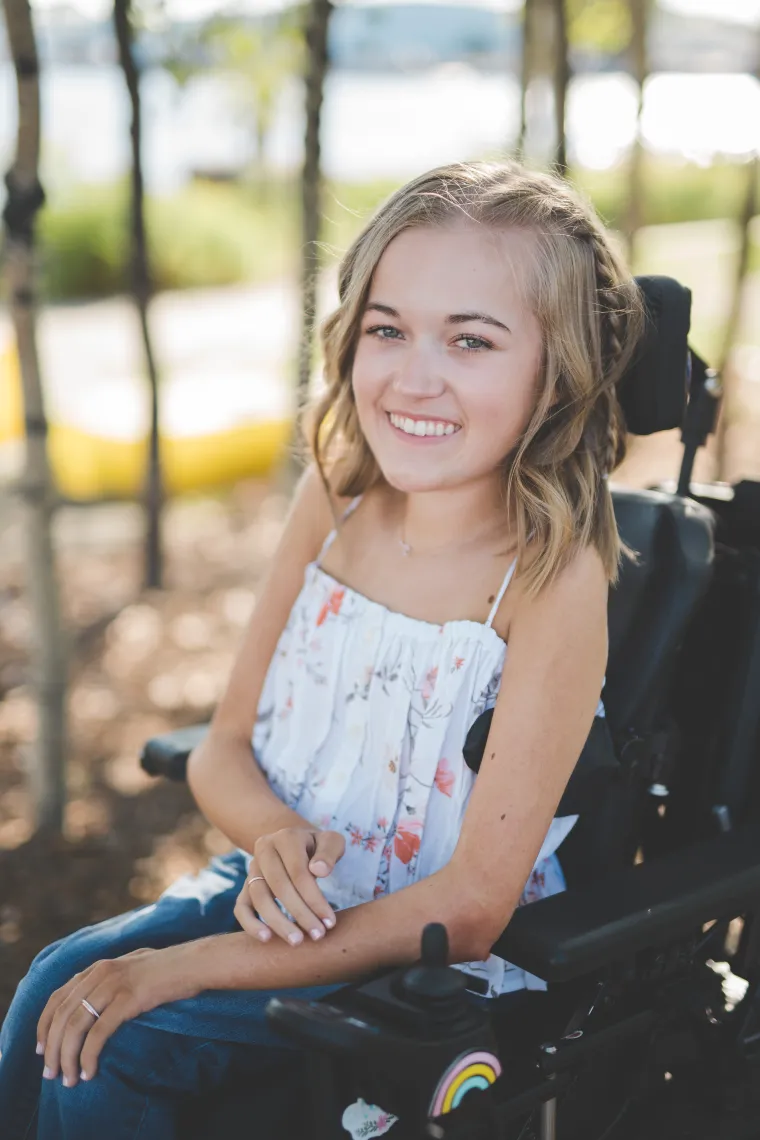 Woman in her power wheelchair with blonde hair wearing a white shirt smiling at the camera