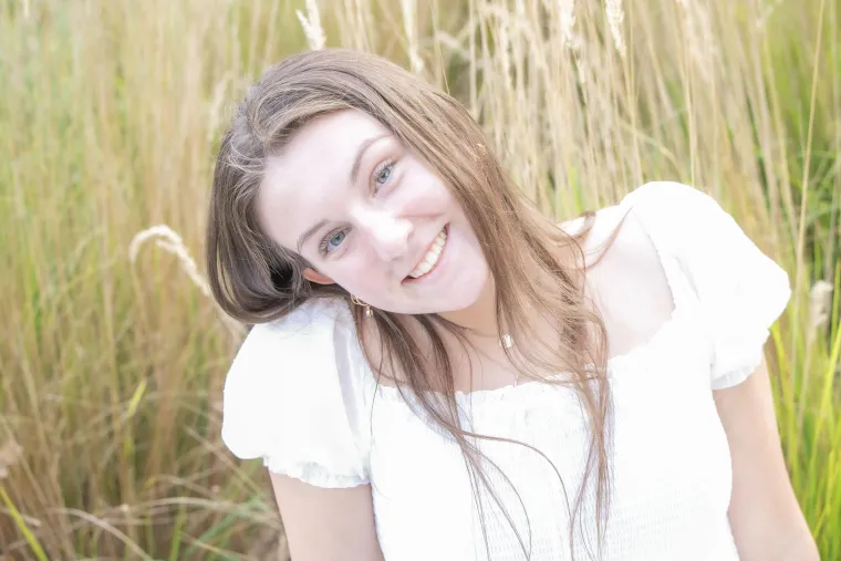 Photo of young woman in front of a field, wearing a white shirt with long brunette hair smiling at the camera