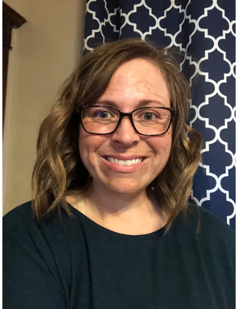 Photo of woman with brunette shoulder length hair wearing a dark blue shirt and black rimmed glasses, and she is smiling at the camera