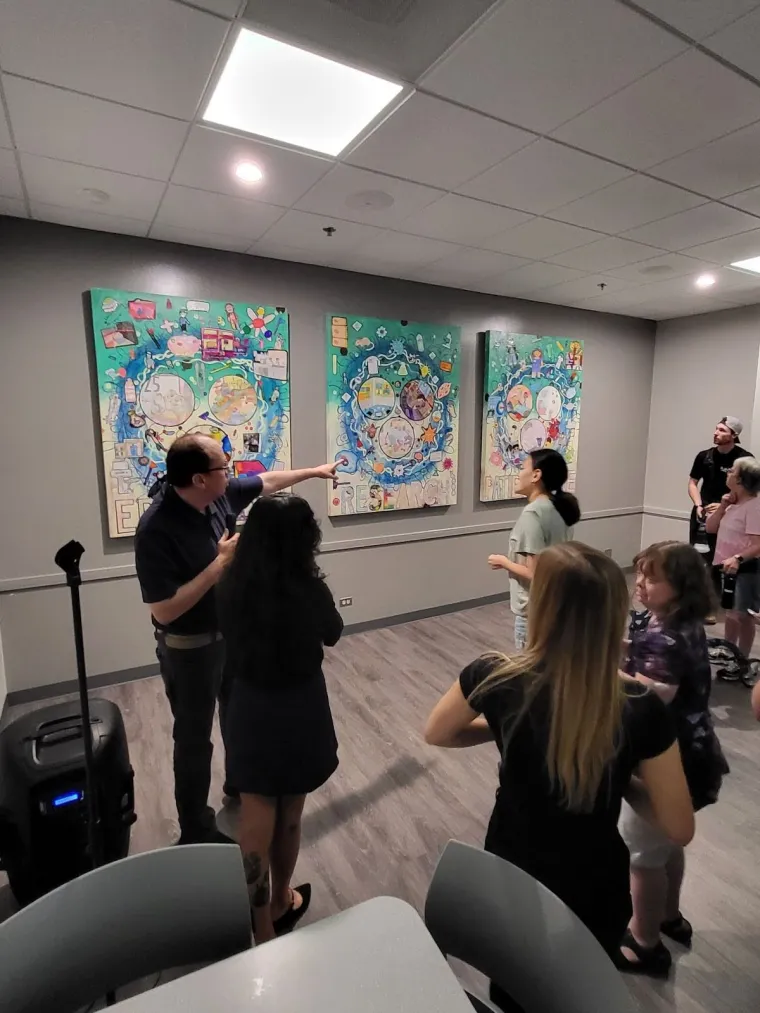 Jack points out his contribution to the mural. He is standing in front of a crowd and pointing towards the "Patient Care" panel.