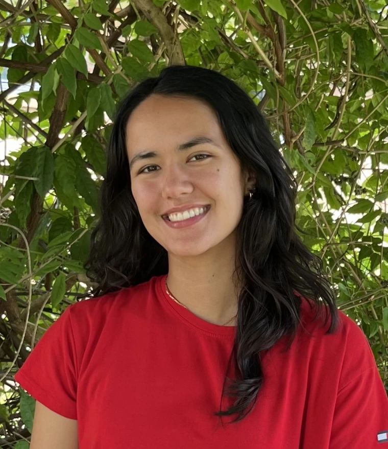 In the center of this image is me, Jeciana, an individual with light colored skin and dark black hair that falls just below my shoulder. In the picture, I am wearing a red shirt and am smiling in front of a large green plant.