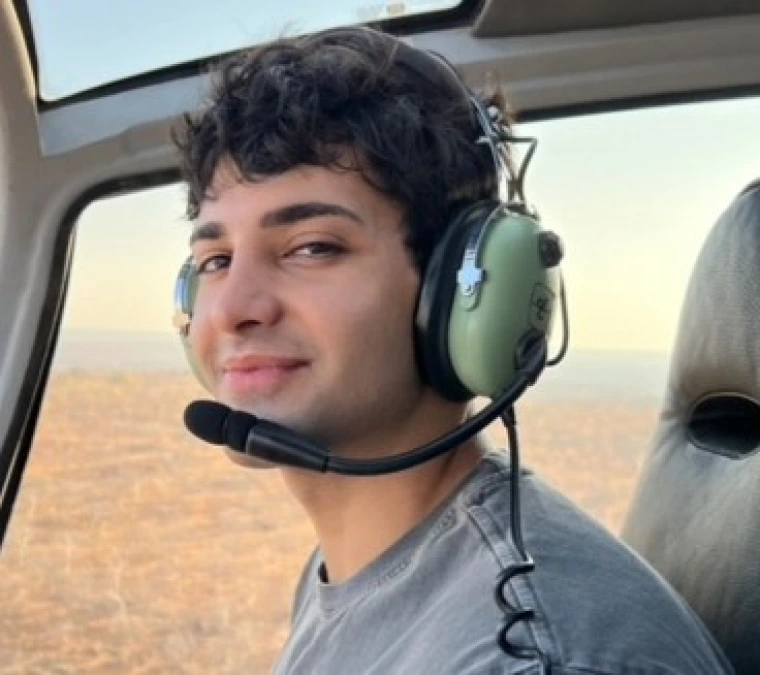 Photo of young man wearing a blue grey long sleeve shirt, has pilot headphone on and is in a helicopter. He has brown curly short hair and is smiling at the camera