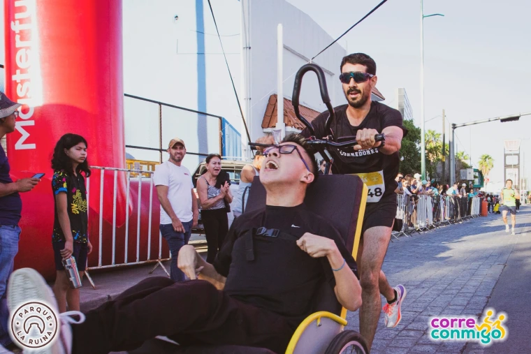 A smiling racer in a wheelchair, helped by a supporter, crosses the finish line