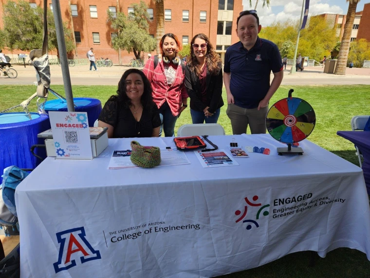 U of A College of Engineering ENGAGED representatives sitting at their table. The table has fliers and a spinning pinwheel with various prizes.
