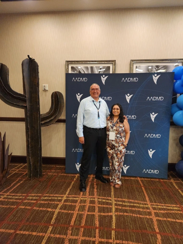 Jim Warne and Celina Urquidez pose in front of a banner with the logo of the AADMD One Voice Conference.