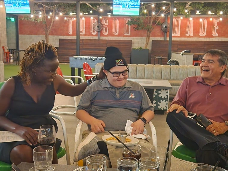Mai, Gabe, and Gabe’s father smile as Gabe puts on a hat with the Tanzanian flag. 