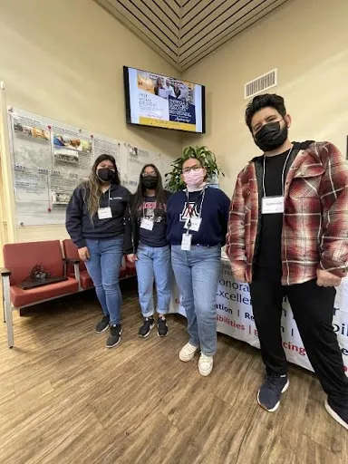 : The four fellows standing in front of a table that has a tablecloth that says Sonoran Center for Excellence in Disabilities. They are in the waiting room of a clinic. Left to right for fellows: Bianca Aguilar is wearing a blue sweater with jeans and black tennis shoes, Sarah Robles is wearing a blue sweatshirt with Arizona is big red letters with blue jeans and black tennis shoes, Paulette Nevarez is wearing a blue sweater with an A for University of Arizona in the middle with blue jeans and white shoes