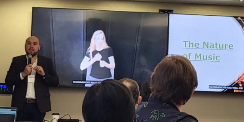 Person wearing a suit talking into a microphone with a person signing ASL and a powerpoint deck on a display monitor in the background that says "The Nature of Music"