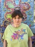Photo of Anne, an older woman with short brown hair, curtain bangs, and pale skin. She is wearing a light green shirt with the image of a colorful flower on it and standing in front of a mosaic mural of a flower.