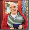 Image of Jimmy, an old man with white hair wearing a red jacket over a green and white striped collared top smiling in front of a red-framed window.