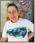 Image of Karen, a woman with brown, pulled-back hair wearing a white shirt with a picture of Orca whales on it, smiling in front of a mosaic mural.