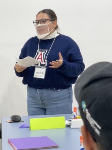 Paulette Nevarez, a woman wearing glasses, a clear plastic face mask, blue UArizona sweatshirt, and jeans holding a piece of paper and talking to project participants