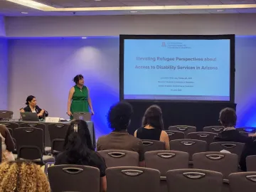 Jacy Farkas and Julie Armin in front of a projector screen in a conference room delivering their presentation Elevating Refugee Perspectives about Access to Disability Services in Arizona. 