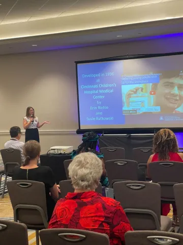 Heather Deitrich delivers her portion of the presentation. She is standing next to a projector screen and in front of a crowd. 