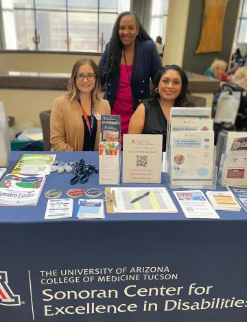 Elizabeth Jeffry-Franco and Celina Urquidez sitting at the Sonoran Center exhibitor table at AACD. Lisa Ayo stands behind them. On the table are many brochures and pamphlets, as well as items for attendees like stickers and hand sanitizer bottles with Sonoran Center branding. 