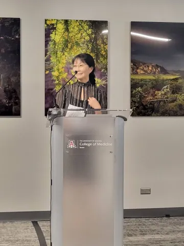 ArtWorks director Yumi Shirai giving an introductory speech behind a podium. Posters of trees and natural landscapes are behind her. 