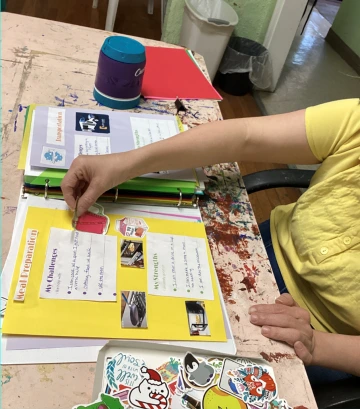 A person in a yellow shirt placing a cut-out picture into the margins of a scrapbook. The open pages contain sections labeled "my challenges" and "my strengths".