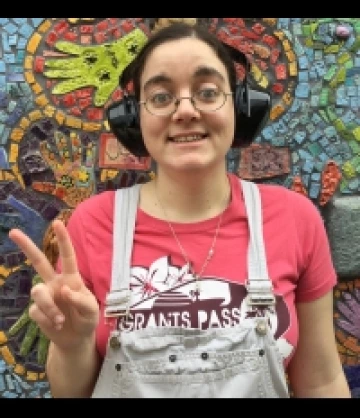 Photo of Abby, a woman with pulled-back brown hair, pale skin, wearing glasses, black headphones, a pink shirt, and a white apron. She is smiling and throwing a peace sign.